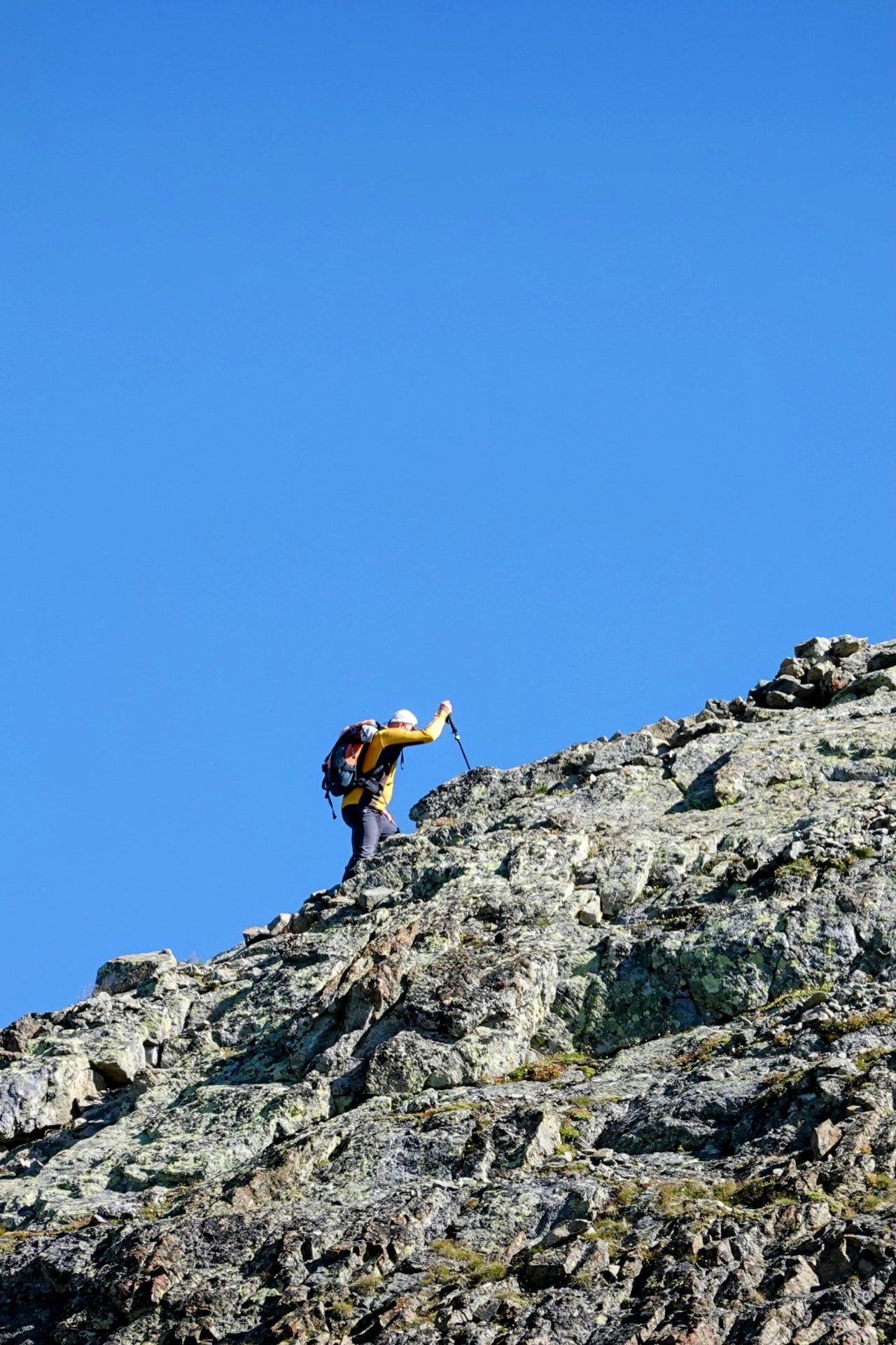 Bergsteiger steigt mit Stock über einen felsigen Grat unter tiefblauem Himmel