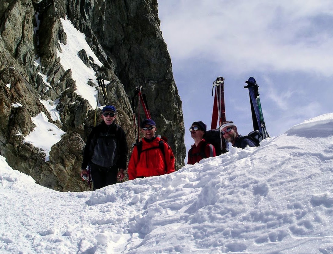 Vier Tourengänger mit Ski am Rucksack in einer Scharte vor dunkler Felswand