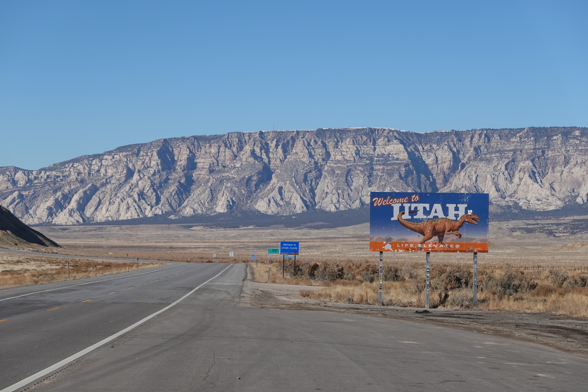 Leere Highway-Strecke mit Schild »Welcome to Utah« vor einer Felskette