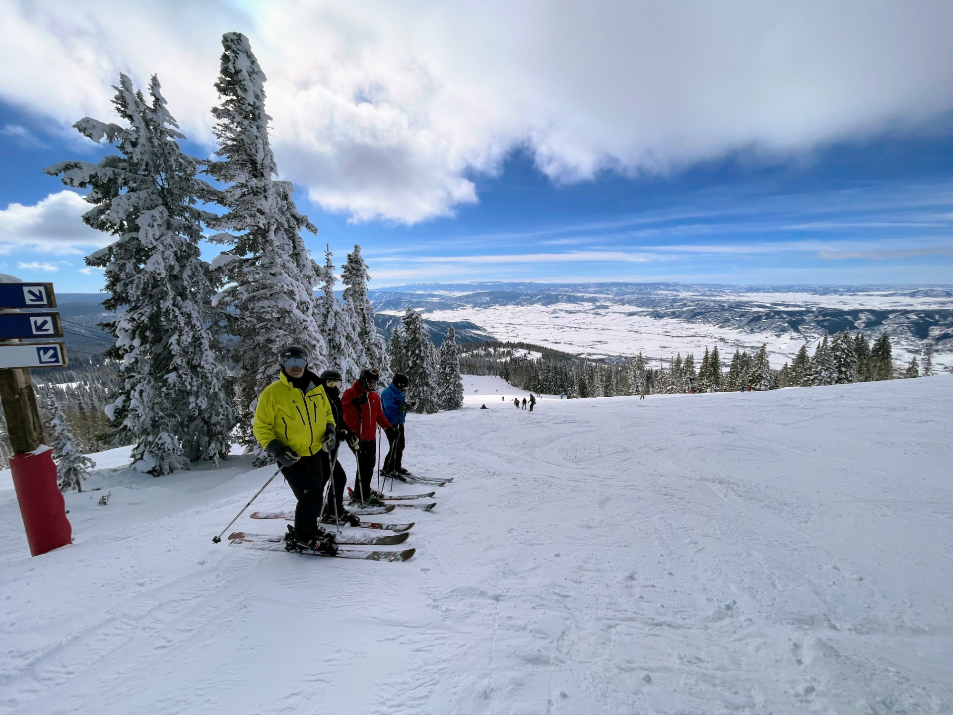 Vier Skifahrer stehen nebeneinander am Pistenrand vor verschneiten Tannen